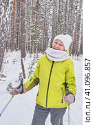 Happy smiling senior woman in yellow jacket, white knitted hat and scarf with ski poles over background of winter snowy forest. Стоковое фото, фотограф Ирина Кононова / Фотобанк Лори