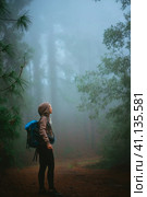 Female traveler wearing hood on the road in the mysterious foggy pine forest. Santo Antao Island, Cape Verde. Стоковое фото, фотограф Igor Tichonow / Фотобанк Лори