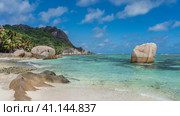 Granite boulders on a sandy beach. Cliffs with smoothed steep slopes rise above the turquoise ocean. Green tropical vegetation on the hill. Clouds in the blue sky. Seychelles. La Digue. Стоковое фото, фотограф Вера Ивановна Кубарева / Фотобанк Лори