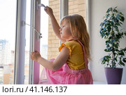 Safety at home for small children. a lock on the window protects children from opening the window. The girl 4 years old tries to open the window standing on the windowsill on a high floor. Стоковое фото, фотограф Ирина Старикова / Фотобанк Лори