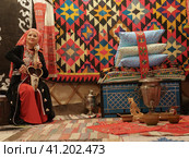 Moscow, Russia - September 15, 2019: Traditional atmosphere, bright home furniture, decoration in Bashkiria. Old lifestyles, fashion at home. Floral pattern of carpet. Family play squeezebox and kubyz. Редакционное фото, фотограф YuryKara / Фотобанк Лори