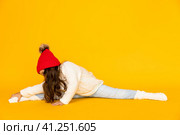 Funny child girl in a white sweatshirt and red knitted hat with pompom doing twine on a yellow background. The concept of children's emotions, joy and delight. Advertising clothing and sales. Стоковое фото, фотограф Ирина Михайличенко / Фотобанк Лори