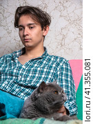 A teenager is sitting on the couch and stroking a gray cat sitting next to him. Стоковое фото, фотограф Ярославский Максим / Фотобанк Лори