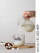 A hand pours milk from a jug into a glass cup with cocoa, chocolate bomb. Making hot chocolate. Vertical frame, light background. Стоковое фото, фотограф Bogdanova Mariia / Фотобанк Лори