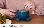 A Hand is Mixing the Milk Foam of Latte. Person Stirring Cappuccino with Steel Spoon. Hot Coffee in a Dark Blue Ceramic Cup on a Wooden Table. Modern Coffee shop, Cafeteria. Thick Froth. Close-up View. Стоковое видео, видеограф Марина Демидюк / Фотобанк Лори