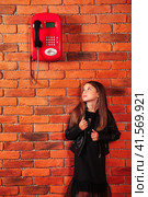 Lovely little kid girl in leather jacket looking up on old red payphone at brick wall, waiting call. Cute child lady model posing near telephone. Support communication concept. Copy ad text space. Стоковое фото, фотограф Aleksandr Golubev / Фотобанк Лори