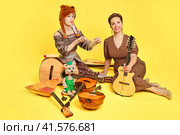 Women musicians in dresses with musical instruments on a yellow studio background. Happy artists with stringed musical instruments with smiles on faces. Стоковое фото, фотограф Андрей Журавлев / Фотобанк Лори