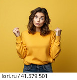 A young, pretty, attractive and thoughtful European girl of 20 years old in a gray casual T-shirt, arms spread apart, isolated on a uniform yellow background. Created by AI. Стоковое AI-изображение, AI-художник Ярославский Максим / Фотобанк Лори
