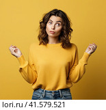A young, pensive, insecure, rather attractive European girl of 20 years in a yellow casual blouse, spreads her hands questioningly, looking away. Created by AI. Стоковое AI-изображение, AI-художник Ярославский Максим / Фотобанк Лори