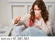 A sick woman blows her nose into a napkin with a thermometer in her hand, fever and high temperature. Adult ill woman with a red scarf sitting on a home bed in a white bedroom, female aged 35 years. Стоковое фото, фотограф Андрей Журавлев / Фотобанк Лори
