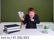 A cunning boy with money in his hands at a desk in a school classroom. US dollars in junior's hand on a blackboard background, copy space for text. Стоковое фото, фотограф Андрей Журавлев / Фотобанк Лори
