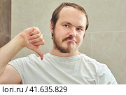 A grown man with half a beard sadly points a finger down. The concept of grooming and problems with appearance when isolated from the coronavirus epidemic. Стоковое фото, фотограф Андрей Журавлев / Фотобанк Лори