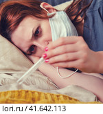 A woman with red eyes, sick with the flu, looks sadly at the thermometer. The patient lies on a bed in a medical mask. Стоковое фото, фотограф Андрей Журавлев / Фотобанк Лори