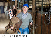 Elderly female rancher walking in stable with saddle in hands. Стоковое фото, фотограф Яков Филимонов / Фотобанк Лори