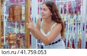 Interested young woman viewing various copper utensils placed on display case of souvenir store. Стоковое видео, видеограф Яков Филимонов / Фотобанк Лори