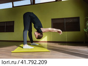 Молодая женщина выполняет упражнение Дандаямана Мудрасана, наклон вперед с вывернутыми руками, занимается йогой в черных легинсах и футболке на коврике в фитнес-центре. Стоковое фото, фотограф Сергей Чайко / Фотобанк Лори