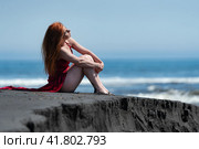 Woman in red long dress with raised hem and bare legs sitting on black sand on beach, hugging knees. Стоковое фото, фотограф А. А. Пирагис / Фотобанк Лори