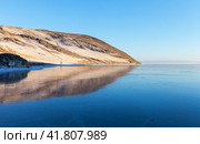 Scenic winter landscape of frozen Baikal Lake on cold January day at sunset. View of snow-capped coastal hills in light of setting sun and icy bay with transparent glass surface of blue ice. Winter ice travel. Стоковое фото, фотограф Виктория Катьянова / Фотобанк Лори
