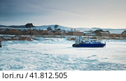 A parked hovercraft in blue next to brown wooden houses against a backdrop of mountains. Winter, shore of Lake Baikal, Olkhon Island. Ice and snow all around. Rescue airboat. Siberia, Russia. (2022 год). Редакционное видео, видеограф Даниил Хабаров / Фотобанк Лори