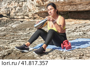 Latin woman, middle-aged, resting, regaining strength, eating, drinking water, after a gym session, burning calories, keeping fit, outdoors by the sea, wearing headphones and smart watch. Стоковое фото, фотограф carlos l vives / Фотобанк Лори