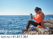 Latin woman, middle-aged, resting, regaining strength, eating, drinking water, after a gym session, burning calories, keeping fit, outdoors by the sea, wearing headphones and smart watch. Стоковое фото, фотограф carlos l vives / Фотобанк Лори