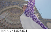 A young woman with long hair in an oriental robe and skullcap stands in the wind near an ancient mosque. Стоковое видео, видеограф Aleksey Kidisyuk / Фотобанк Лори
