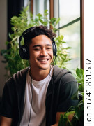 Asian young man listening to music with headphones sitting near window with green house plants. Стоковое AI-изображение, AI-художник Наталья Семкина / Фотобанк Лори