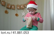 Boy in pajamas, Santa cap eating sweet candy on paper advent calendar with presents background. Little happy child with gift of handmade Christmas calendar on wall. Celebrating at home Xmas tradition. Стоковое видео, видеограф Марина Демидюк / Фотобанк Лори
