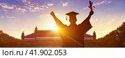 Graduation Celebration at Golden Hour. A silhouetted graduate triumphantly holds a diploma against a breathtaking sunset. Academic achiever, culmination of education. Стоковое AI-изображение, AI-художник Ольга Сухарева / Фотобанк Лори