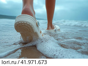 Crocs shod feet of an person on the beach walking along the surf. Стоковое AI-изображение, AI-художник Евгений Харитонов / Фотобанк Лори