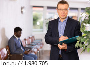 Dispassionate secretary man stands at reception with folder and list of guests and expects visitors. Стоковое фото, фотограф Яков Филимонов / Фотобанк Лори