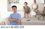 Emotionally tense distressed young guy sitting in home kitchen against blurred background of angry mother and sister standing in back and expressing dissatisfaction. Concept of family problems. Стоковое видео, видеограф Яков Филимонов / Фотобанк Лори