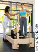 Focused Latin female instructor helping girl practicing pilates stretching exercises on reformer at gym. Individual coach. Стоковое фото, фотограф Яков Филимонов / Фотобанк Лори