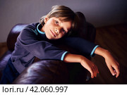 Sad male child is deprived of friends and gadgets as punishment for pranks, he is bored in dimly lit room alone, sitting on brown leather sofa. Стоковое фото, фотограф Владимир Григорьев / Фотобанк Лори