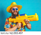 A young man with a beard, sunglasses and a water pistol is smiling, rejoicing at the upcoming vacation. The concept of summer holidays. Стоковое AI-изображение, AI-художник Ярославский Максим / Фотобанк Лори