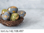 Stylish beautiful Easter eggs with a golden coating of sweat and feathers in a bird's nest of brushwood on a gray background. The concept of happy Easter 2024. Стоковое фото, фотограф Photojuli / Фотобанк Лори