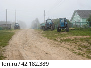 Misty morning in the countryside. View of a deserted village street. Old tractors on the side of a dirt road. Foggy rural landscape. Wooden houses behind fences. Travel across Russia. Vologda region. Стоковое фото, фотограф Андрей Степанов / Фотобанк Лори