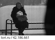 Пожилая женщина сидит на остановке и копается в своей старой сумке (2017 год). Редакционное фото, фотограф Валерий Леонидович / Фотобанк Лори