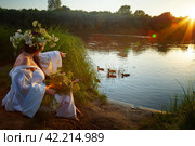 Adult mature brunette woman in a white dress, sundress and a wreath of flowers in summer by the water of river or lake in the evening at sunset. Celebration of the Slavic pagan holiday of Ivan Kupala. Стоковое фото, фотограф Кривошеина Елена Леонидовна / Фотобанк Лори