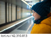 Улыбающийся мальчик в зимней куртке и шапке ждет приближающегося поезда на платформе метро. Стоковое фото, фотограф Яна Проничева / Фотобанк Лори
