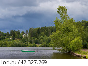 Летний пейзаж с озером в сельской местности. Лодка на воде недалеко от берега. Деревья на берегу озера. Темные тучи в небе перед дождем. Кавголовское озеро, Токсово, Ленинградская область, Россия. Стоковое фото, фотограф Андрей Степанов / Фотобанк Лори