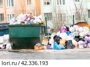 A garbage container site littered with unsorted household waste and a poor anonymous person rummaging through it in search of food. Garbage not removed on time. Overall plan (2024 год). Редакционное фото, фотограф Антон Вахрушев / Фотобанк Лори