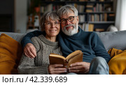 Retired couple resting together on sofa with favourite book. Family leisure and hobby. Стоковое AI-изображение, AI-художник Alexander Tihonovs / Фотобанк Лори