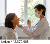 Middle-aged doctor in protective mask examining patient throat and oral cavity. Стоковое фото, фотограф Natalia Gdovskaia / Фотобанк Лори