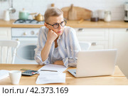 Frustrated senior woman wearing glasses working with documents while sitting in the modern kitchen. Стоковое фото, фотограф Natalia Gdovskaia / Фотобанк Лори