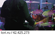 A drummer sitting at a drum kit on a large stage, viewed from behind. The drum kit includes three toms, a hi-hat, and four cymbals. Stage lights and cables are also visible in the image. Стоковое фото, фотограф Воловодов Владимир / Фотобанк Лори