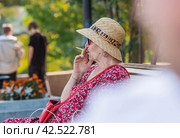 Harm to health. Nicotine addiction. An elderly woman in a straw hat smokes a cigarette on a park bench.Nizhny Novgorod, Russia, August 2023. Редакционное фото, фотограф Photojuli / Фотобанк Лори