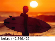 Surfer at Sunset with Surfboard in Silhouette Against Orange Sky and Ocean Waves. Стоковое AI-изображение, AI-художник Юрий Поздников / Фотобанк Лори