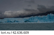 A wall of blue ice with cracks, crevices, and sharp peaks rises above a turquoise glacial lake. Icebergs, melted ice floes float in the water. The mountains are in a fog. Perito Moreno glacier. Стоковое фото, фотограф Вера Ивановна Кубарева / Фотобанк Лори
