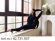 Brunette woman performs yoga exercise at home Parivritta Surya Yantrasana, compass pose, training in sportswear sitting on the windowsill. Стоковое фото, фотограф Сергей Чайко / Фотобанк Лори